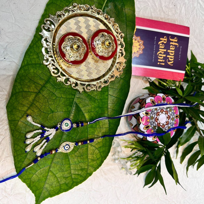 Decorative rakhi set with gold and silver thali, colorful rakhi, and 'Happy Rakhi!' card on a green leaf.